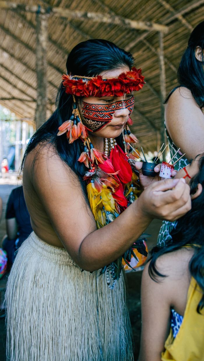 Indigenous woman adorned in tribal attire paints another's face during a cultural ceremony.